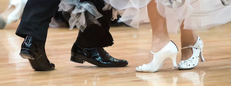 The Height of Heels That Not Harmful 2 The footwear of a couple dancing in heels. The woman is wearing white comfortable shoes with a wide heel, while the man is wearing elevated patent leather shoes.