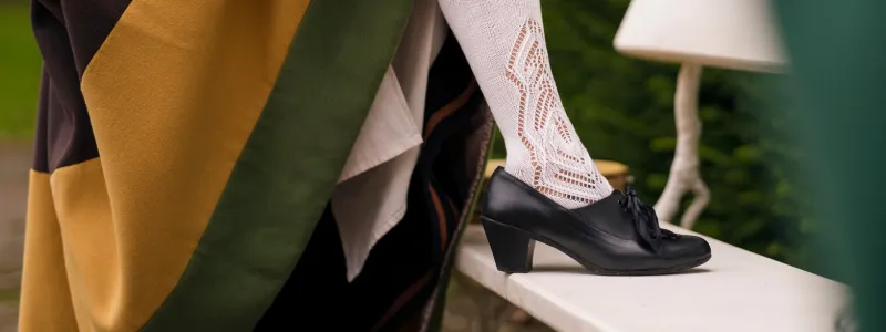 A historical figure in a medieval brown, yellow, and green gown paired with noble black low shoes featuring wide heels.