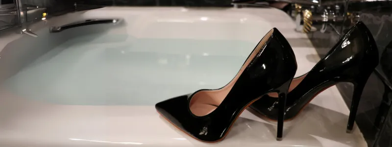 Black patent high heels on the edge of a bathtub filled with water for a bath.