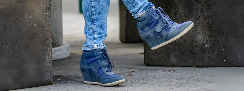 Blue high-top wedge shoes paired with light denim jeans, captured mid-step on a grey urban pavement with concrete structures in the background.