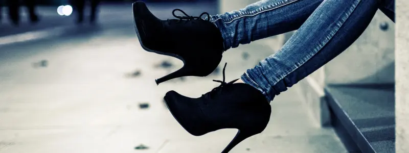 Person wearing black high-heeled ankle boots and blue skinny jeans, sitting on city steps at night.