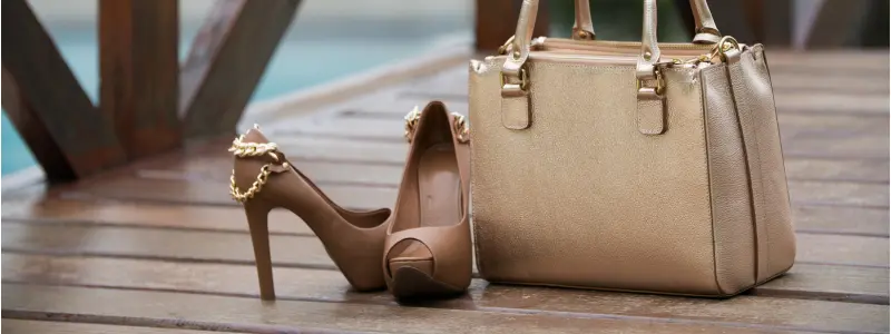 Nude high-heel stilettos placed next to a beige luxury handbag on a wooden deck, representing classy, high-end designer fashion accessories.